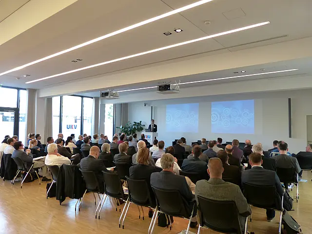 Mehrere Teilnehmer hören dem Referenten zu. Die Aufnahme zeigt die Rücken der Teilnehmenden