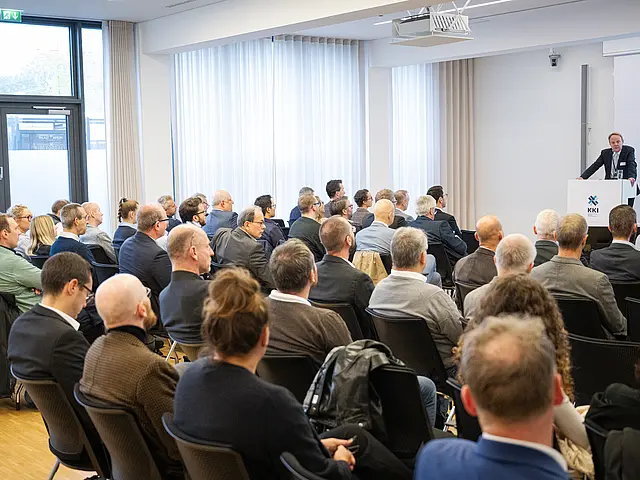 Redner präsentiert. Vorne sieht man viele Teilnehmer von hinten