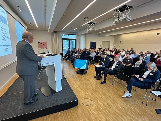 Seitliche Aufnahme des Redners. Rechts sieht man einige Teilnehmer