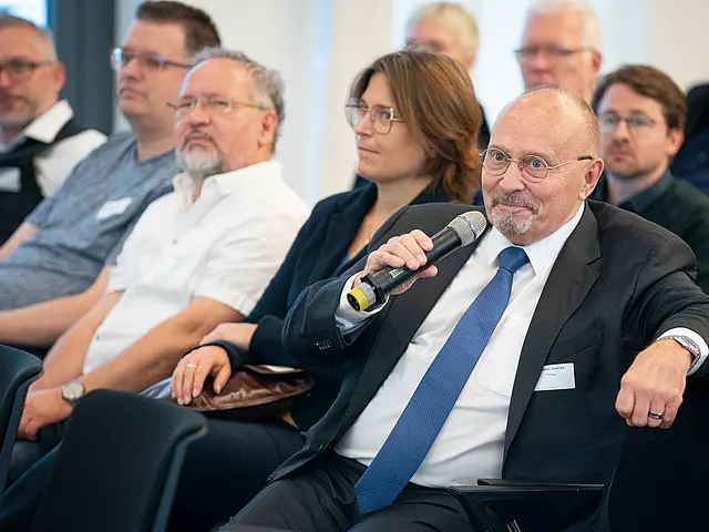 Man hält Mikro und stellt eine Frage. Daneben sitzen mehrere Teilehmer