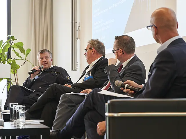 Podiumsdiskussion mit vier Männern. Einer hält das Mikrofon und spricht